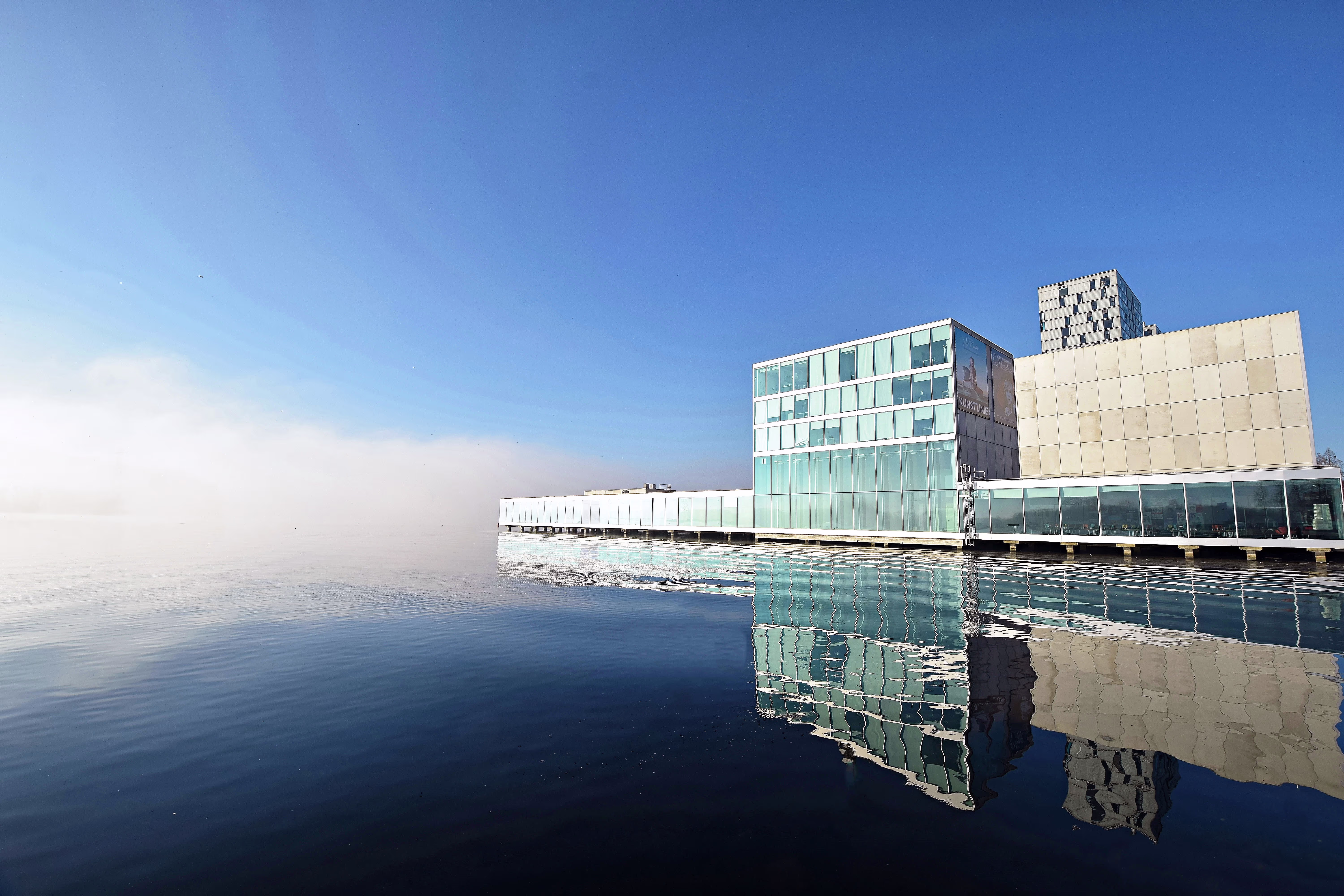 Modern glazen gebouw aan het water met perfecte reflectie op kalm wateroppervlak onder blauwe hemel.