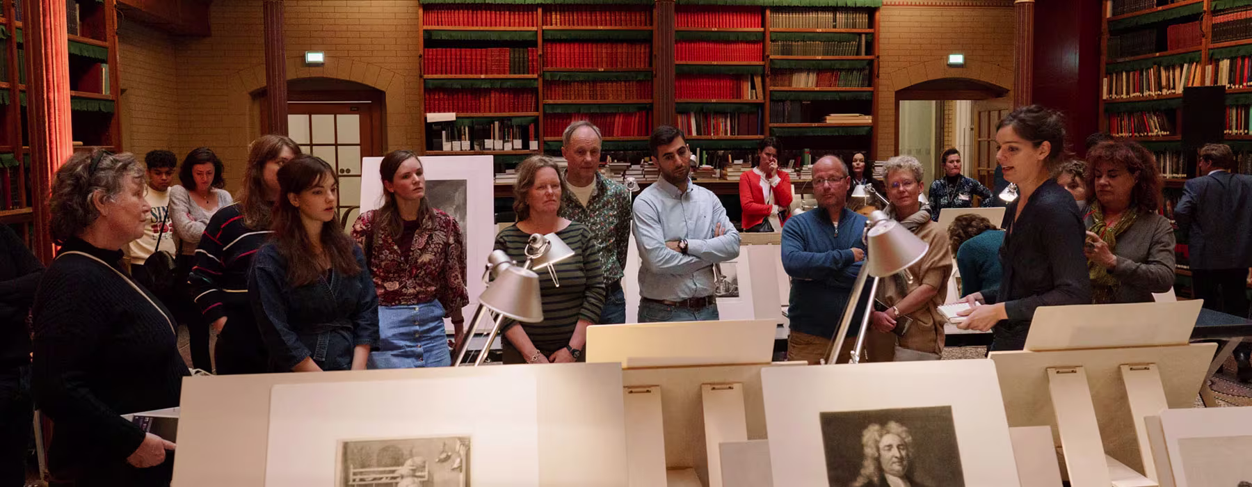 Een groep mensen bekijkt historische kunstwerken en documenten in een bibliotheek met boekenkasten op de achtergrond.