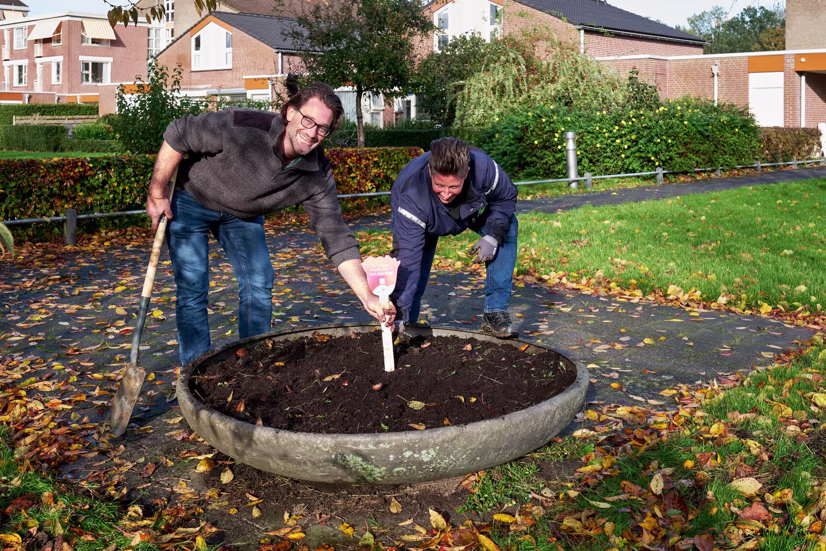 Twee mannen planten samen een boom in een ronde plantenbak in een woonwijk, een met schep en een met gieter.