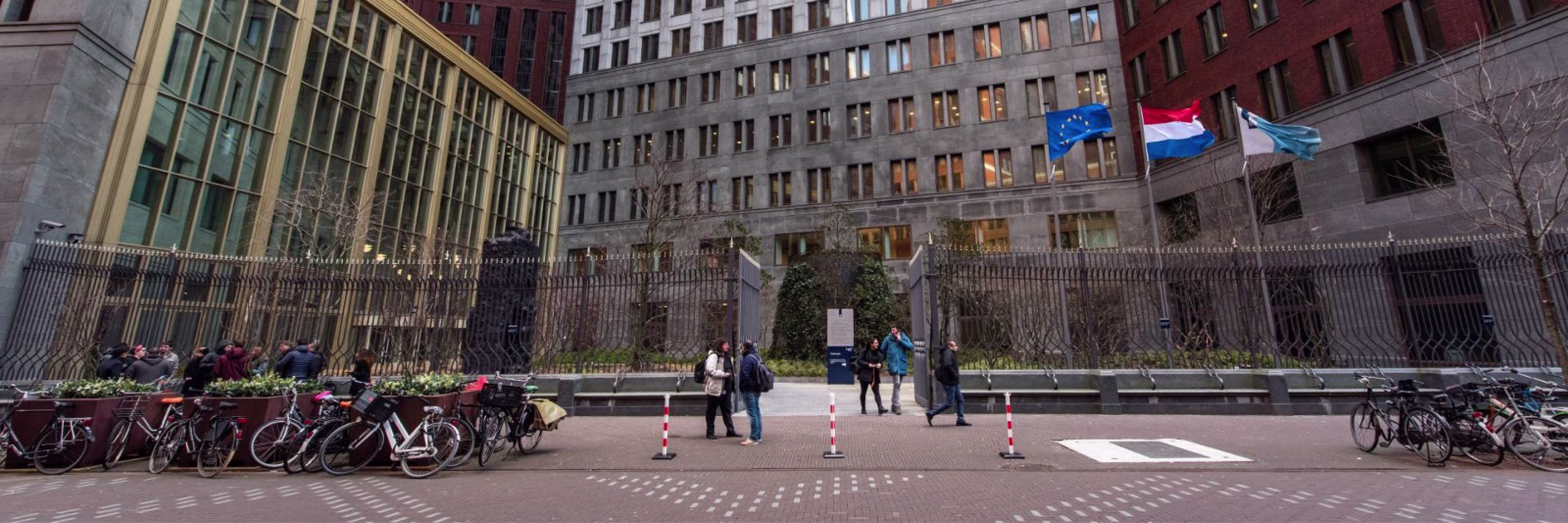 Buitenaanzicht van een modern overheidsgebouw met Nederlandse vlag en fietsers op het plein ervoor.