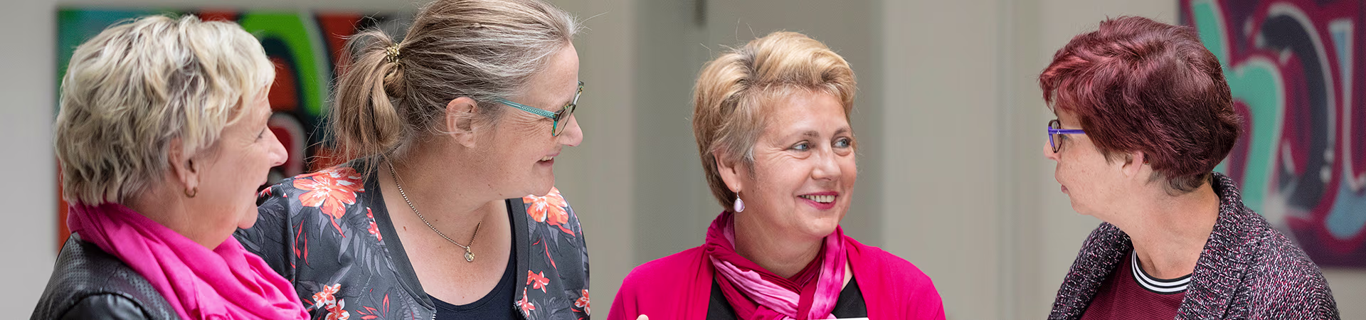 Vier vrouwelijke collega's staan gezellig met elkaar te praten in een moderne kantooromgeving.