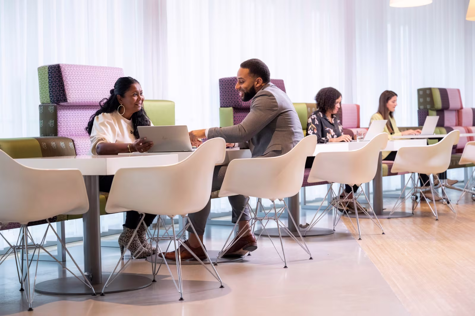Collega's werken samen aan laptops in een moderne, kleurrijke kantooromgeving met witte stoelen en tafels.