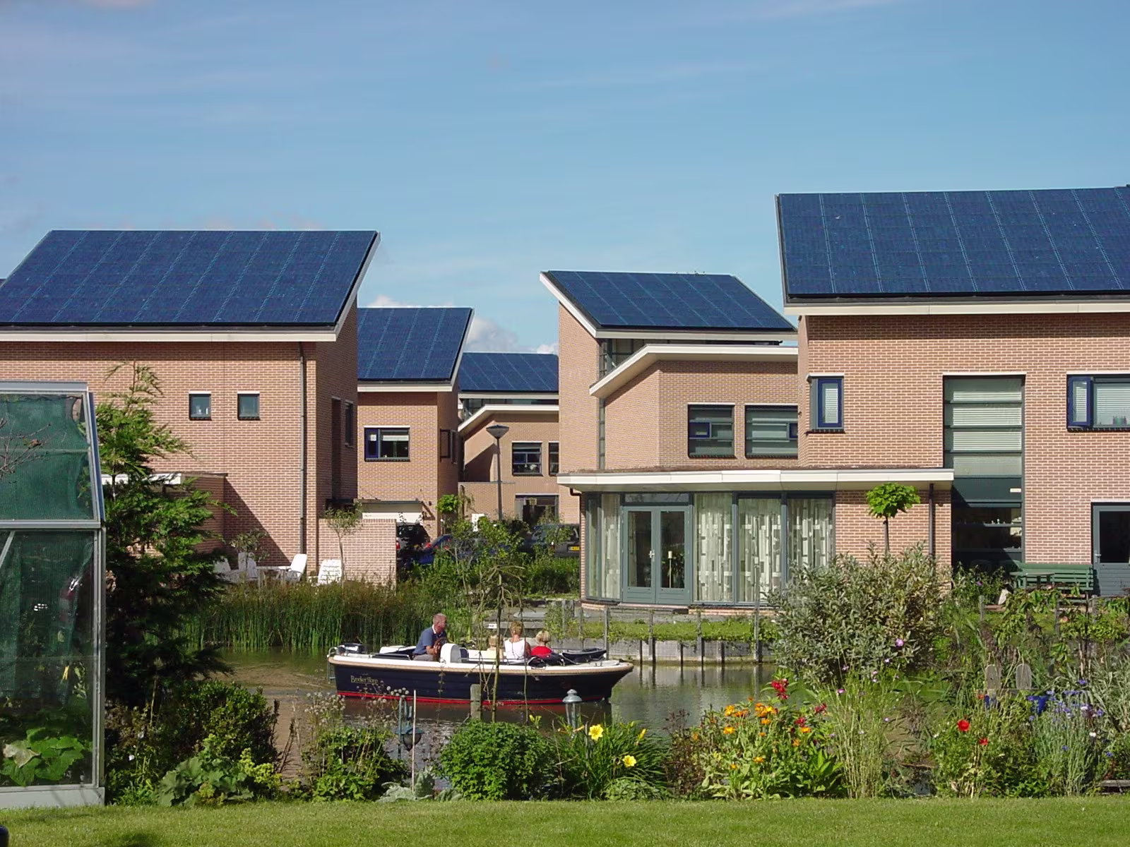 Moderne huizen met zonnepanelen langs een waterweg, met een bootje en groene tuinen in een duurzame woonwijk.