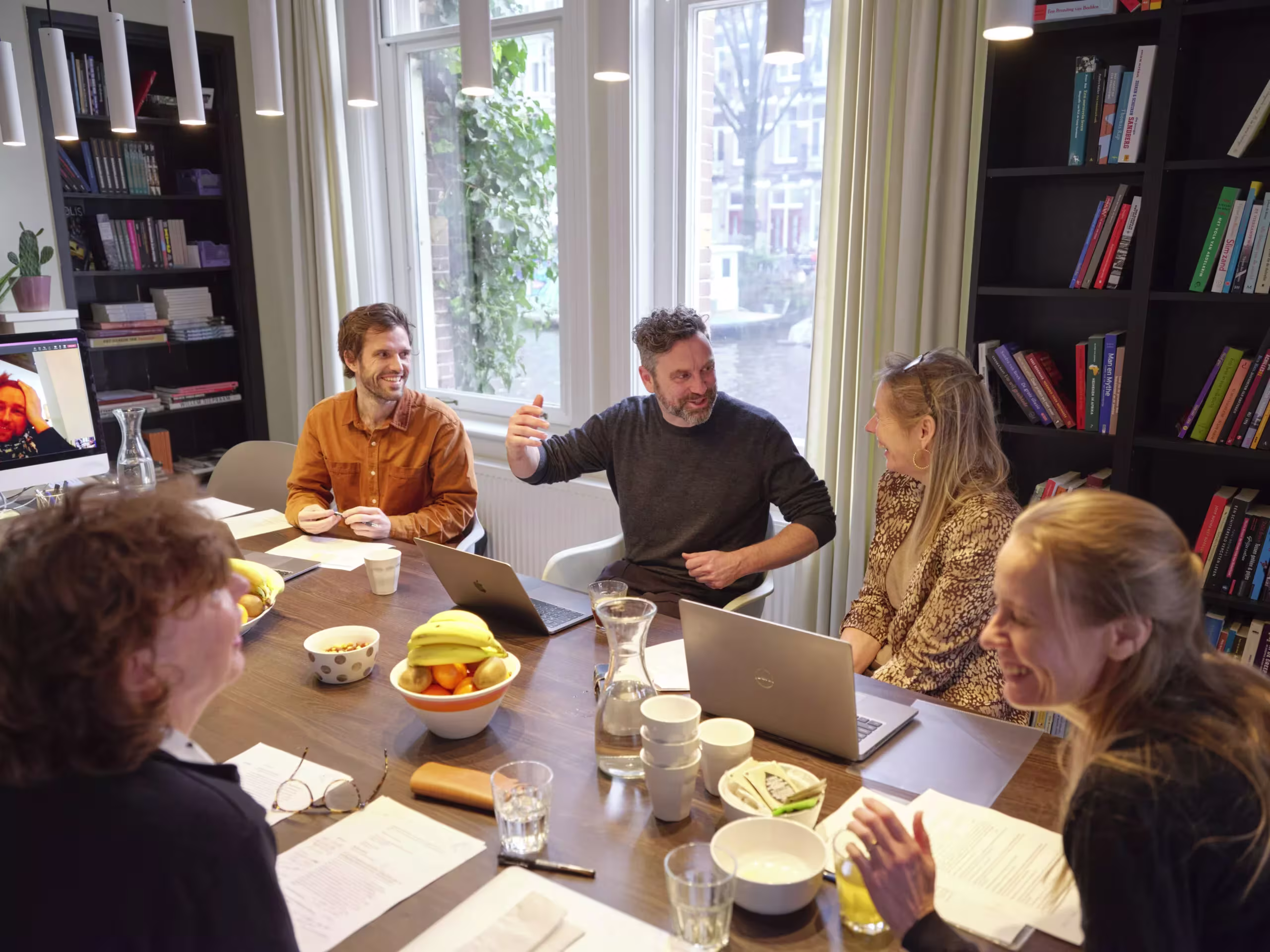 Groep mensen werkt samen aan tafel met laptops, documenten en koffie in kantoor met boekenkasten bij raam.