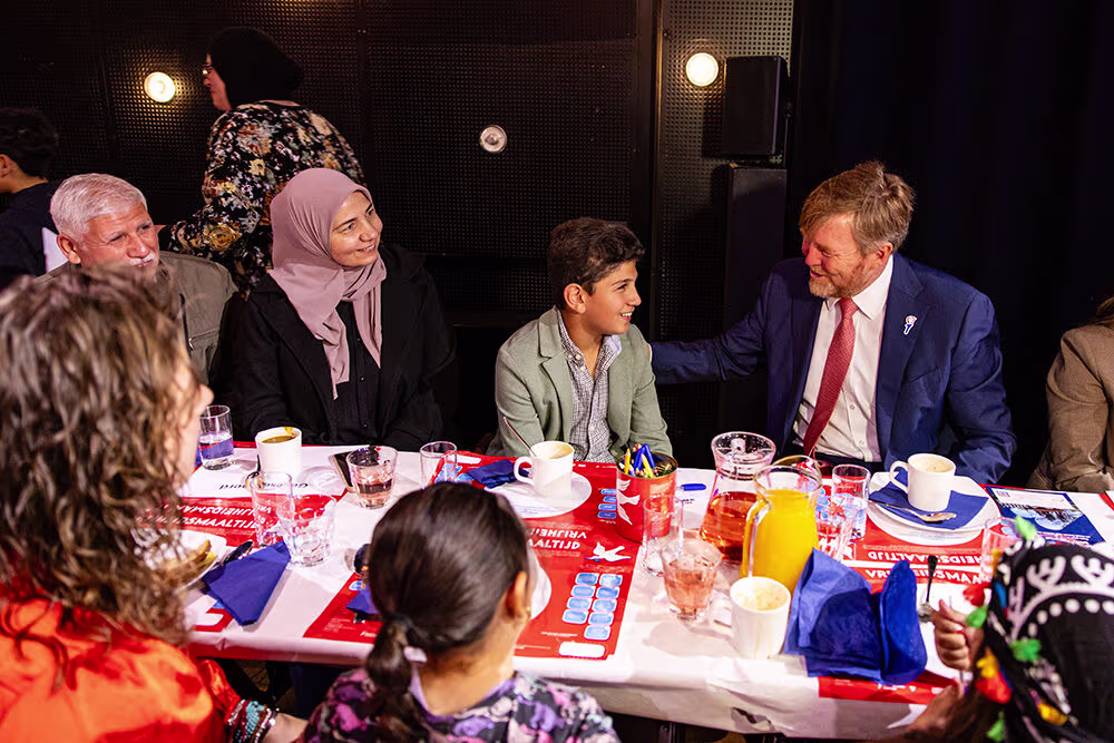 Een man in donkerblauw pak zit aan tafel in gesprek met diverse mensen, waaronder een vrouw met hoofddoek en een jongen.