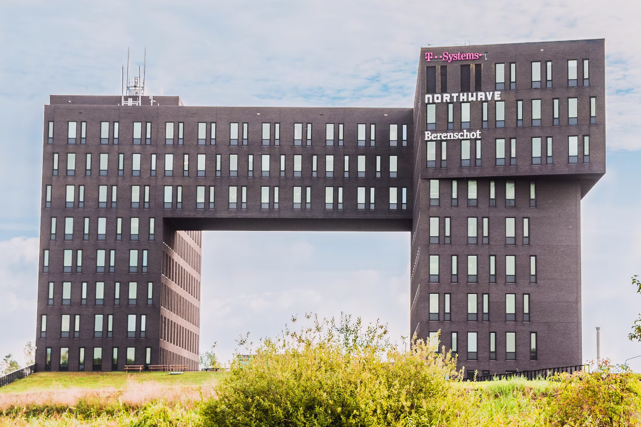 Modern kantoorgebouw van Berenschot met kenmerkende brug-architectuur, omgeven door groen landschap onder bewolkte hemel.