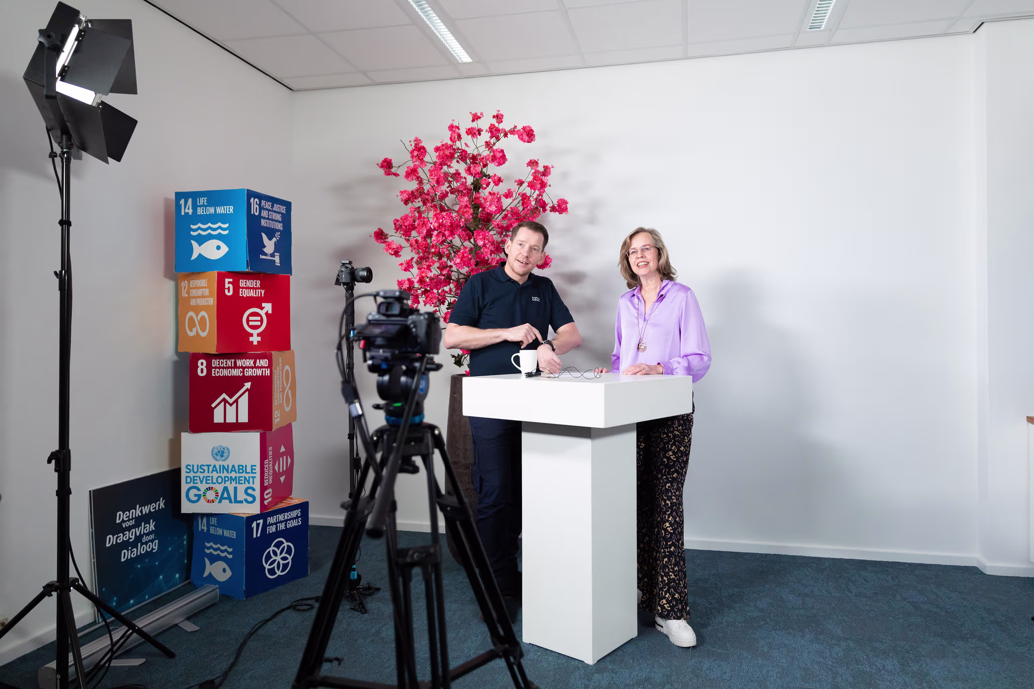 Twee personen staan bij een witte tafel in een studio-opzet met camera's, belichting en SDG-blokken op de achtergrond.