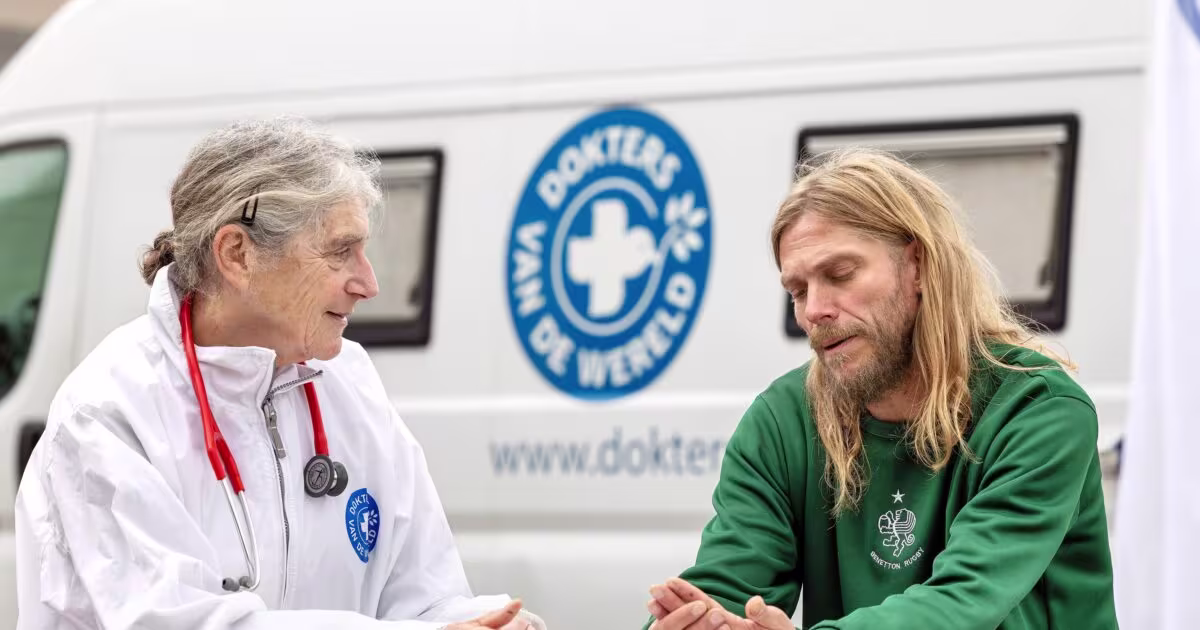 Een arts in witte jas met stethoscoop spreekt met een man in groene trui naast een Dokters van de Wereld ambulance.