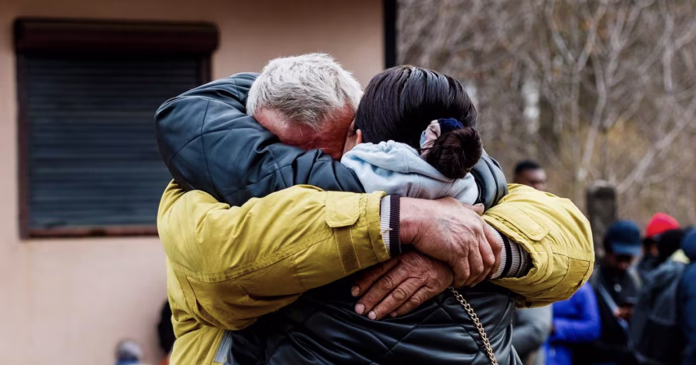 Twee mannen omhelzen elkaar hartelijk, een oudere man met grijs haar en een jongere man, beide in winterjassen.
