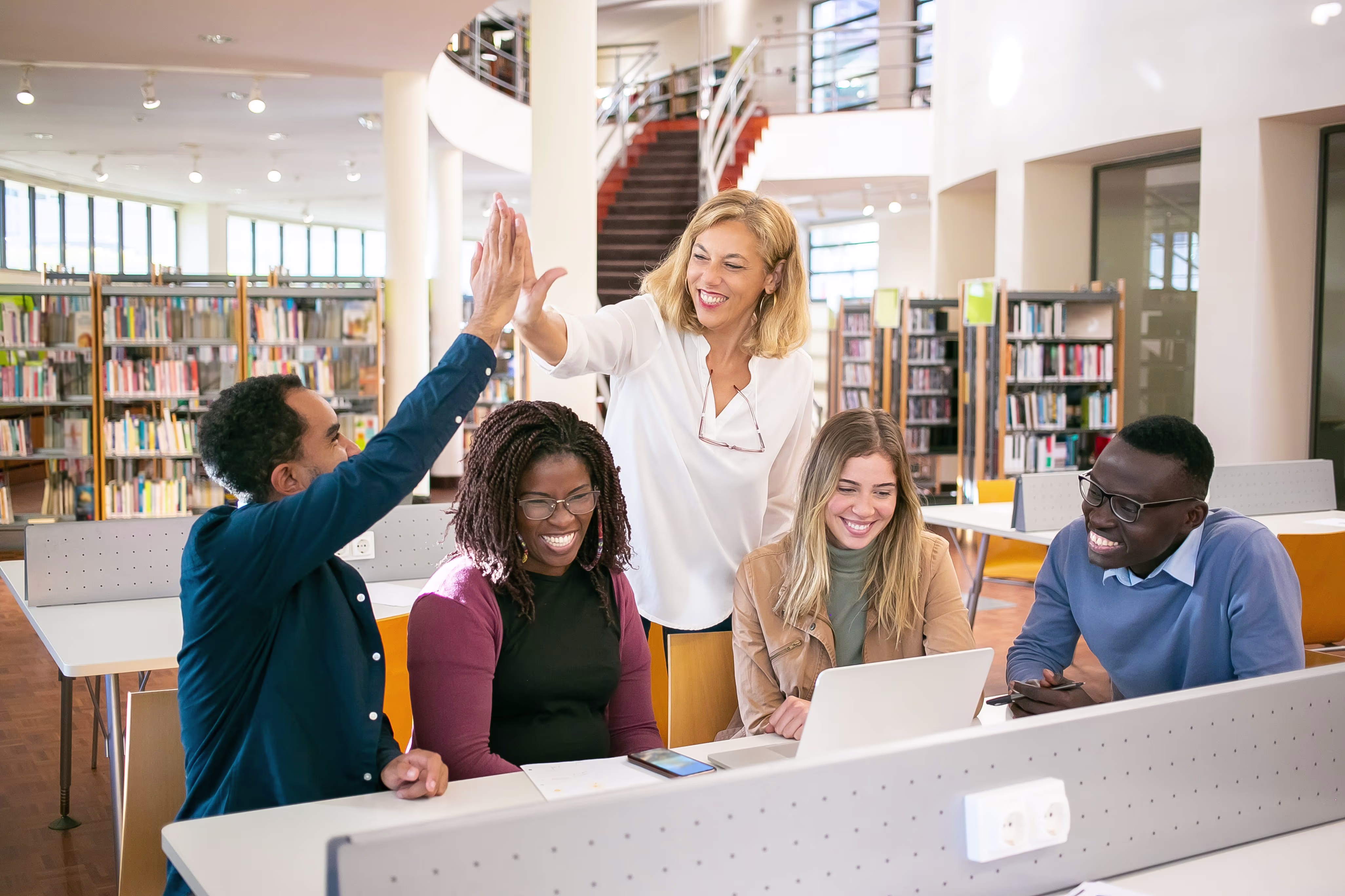 Groep studenten en docent geven high five in moderne bibliotheek van hogeschool, iedereen lacht en kijkt blij.