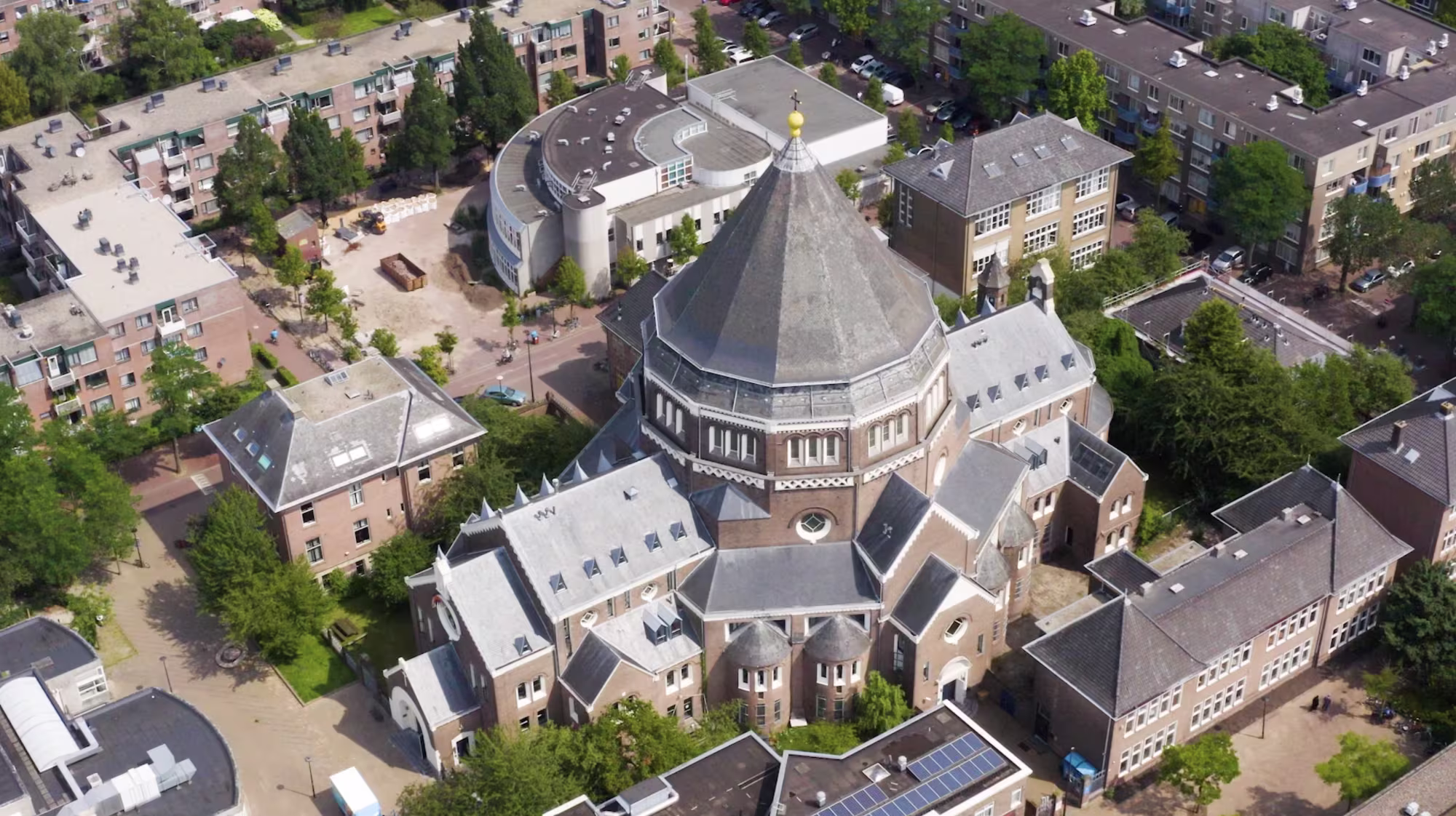 Luchtfoto van een grote kerk met koepeldak en een modern rond gebouw, omgeven door woningen en groen in een woonwijk.