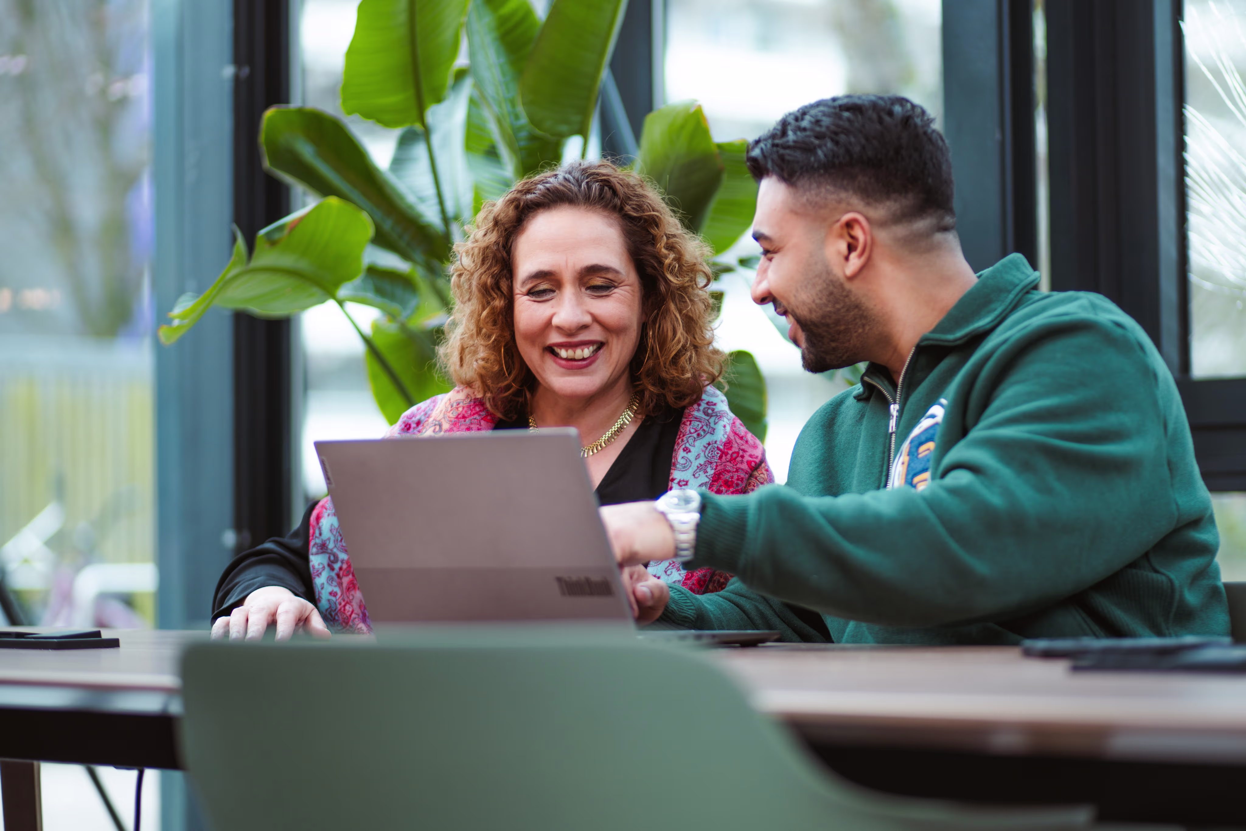 Twee collega's kijken lachend naar een laptop in een moderne werkruimte met veel groen.
