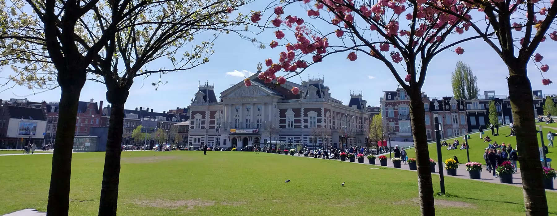 Het Concertgebouw met klassieke architectuur, omgeven door groene parken met bloeiende bomen en bezoekers op een zonnige dag.
