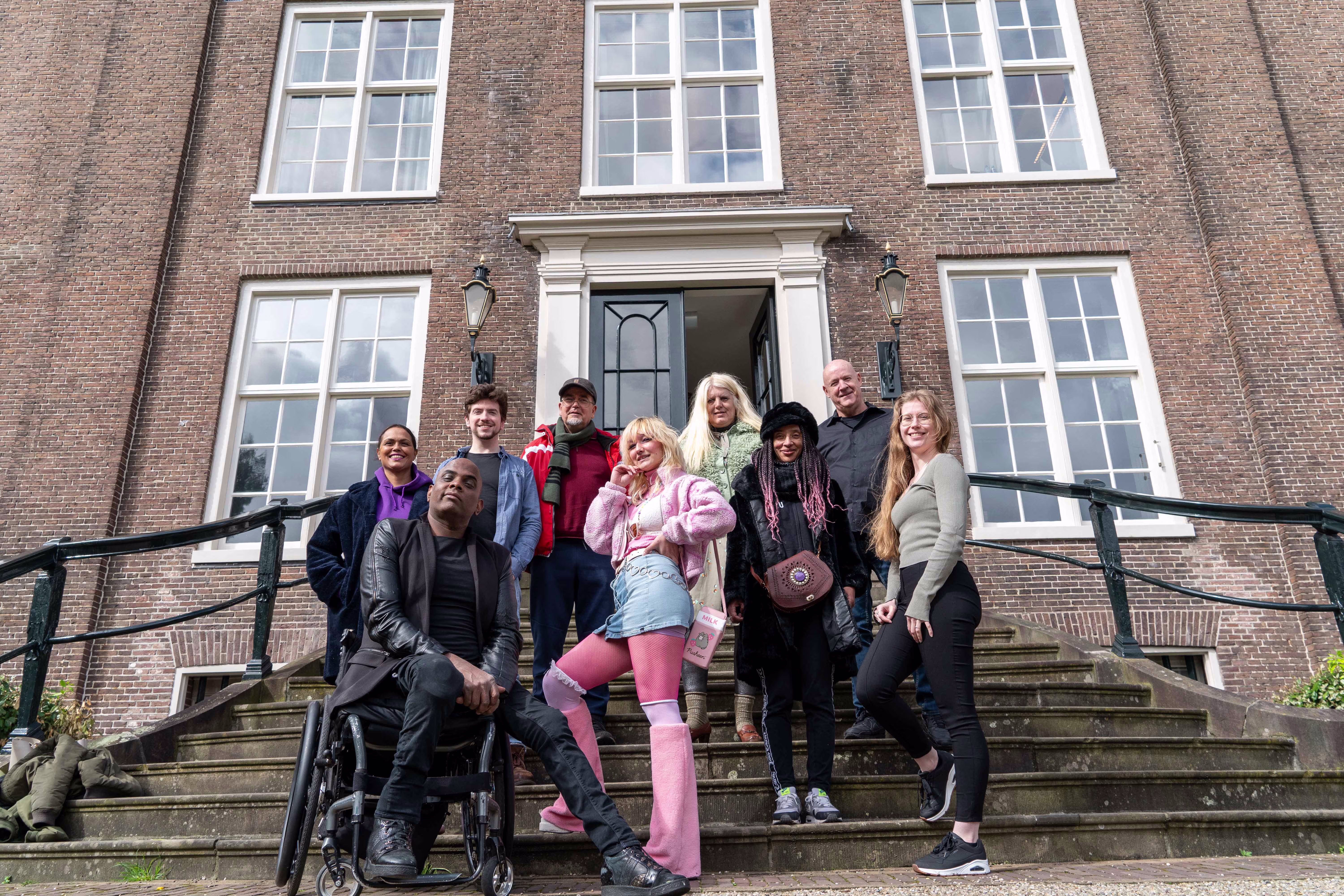 Een diverse groep collega's poseert vrolijk op de trap voor een historisch bakstenen gebouw met witte raamkozijnen.