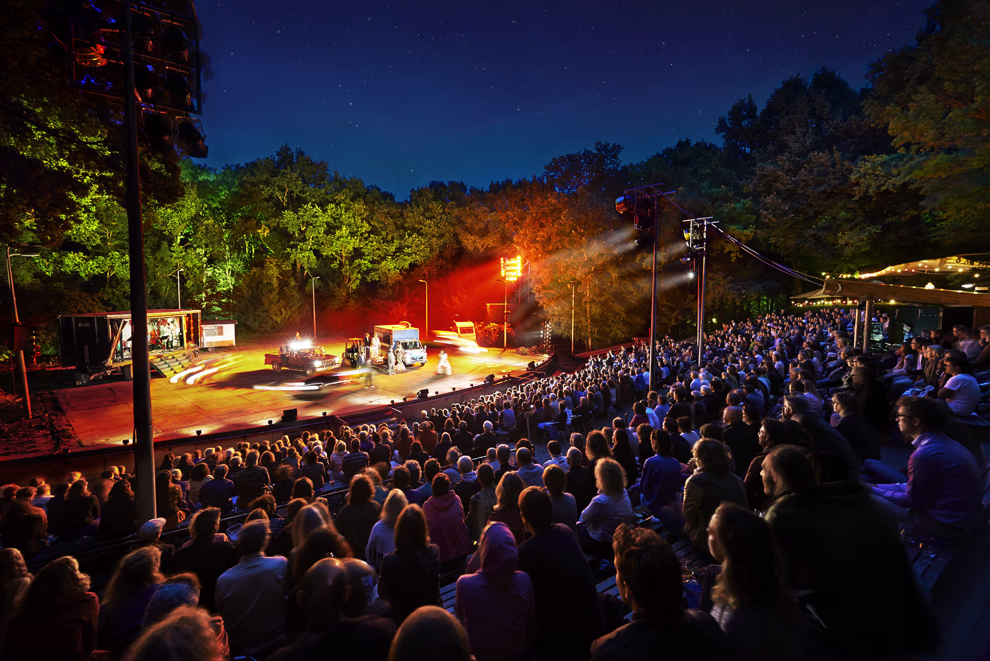 Een avondvoorstelling in een openluchttheater met publiek op tribune en verlichte podium tussen de bomen.