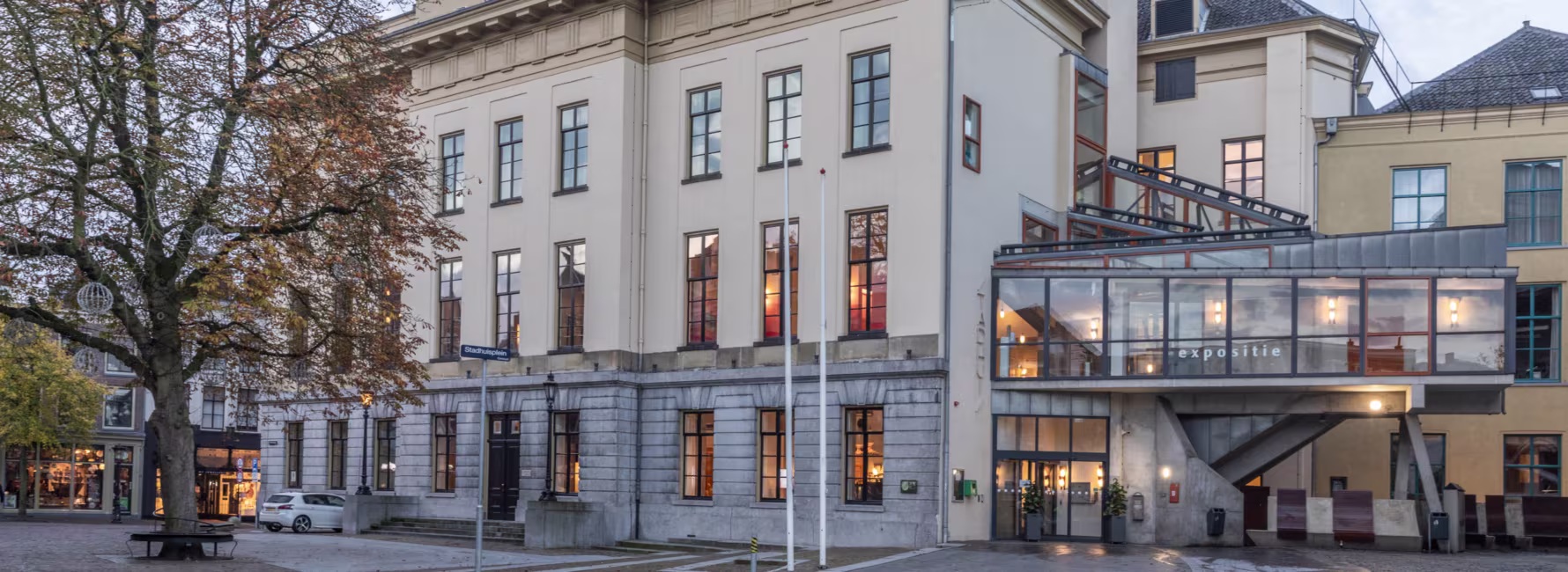 Historisch gemeentehuis van Utrecht met verlichte ramen en moderne glazen aanbouw, omgeven door bomen en andere gebouwen.