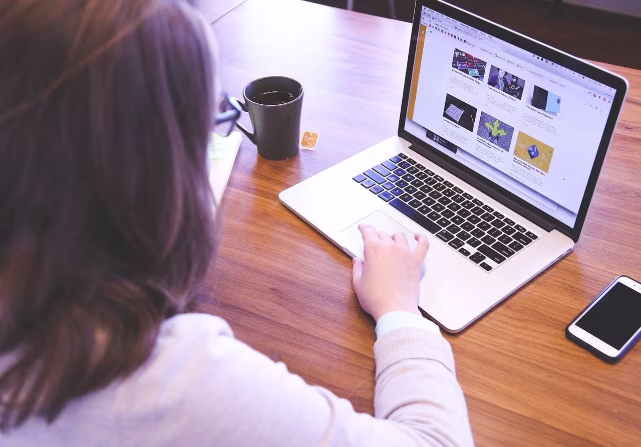 Een vrouw werkt achter een laptop op een houten bureau, met een koffiemok en smartphone naast zich.