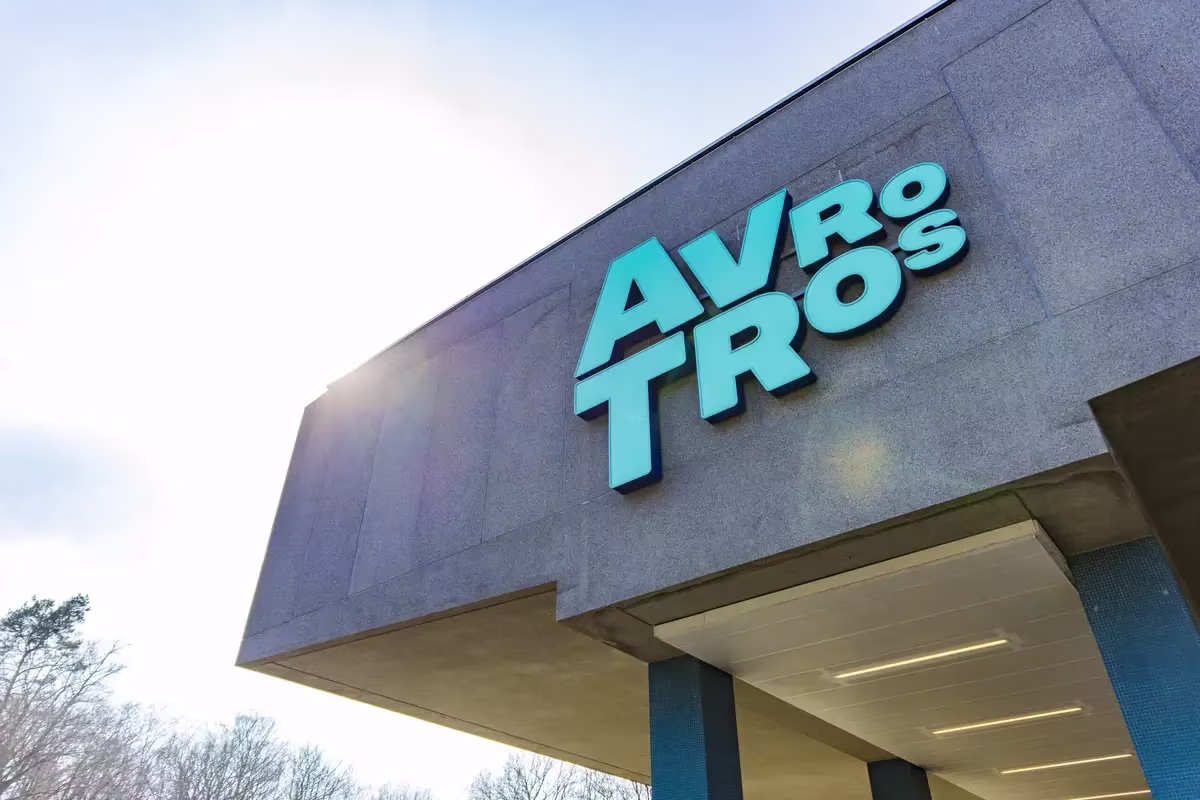Moderne buitenkant van het AVROTROS-gebouw met groot logo op grijze gevel tegen blauwe hemel.