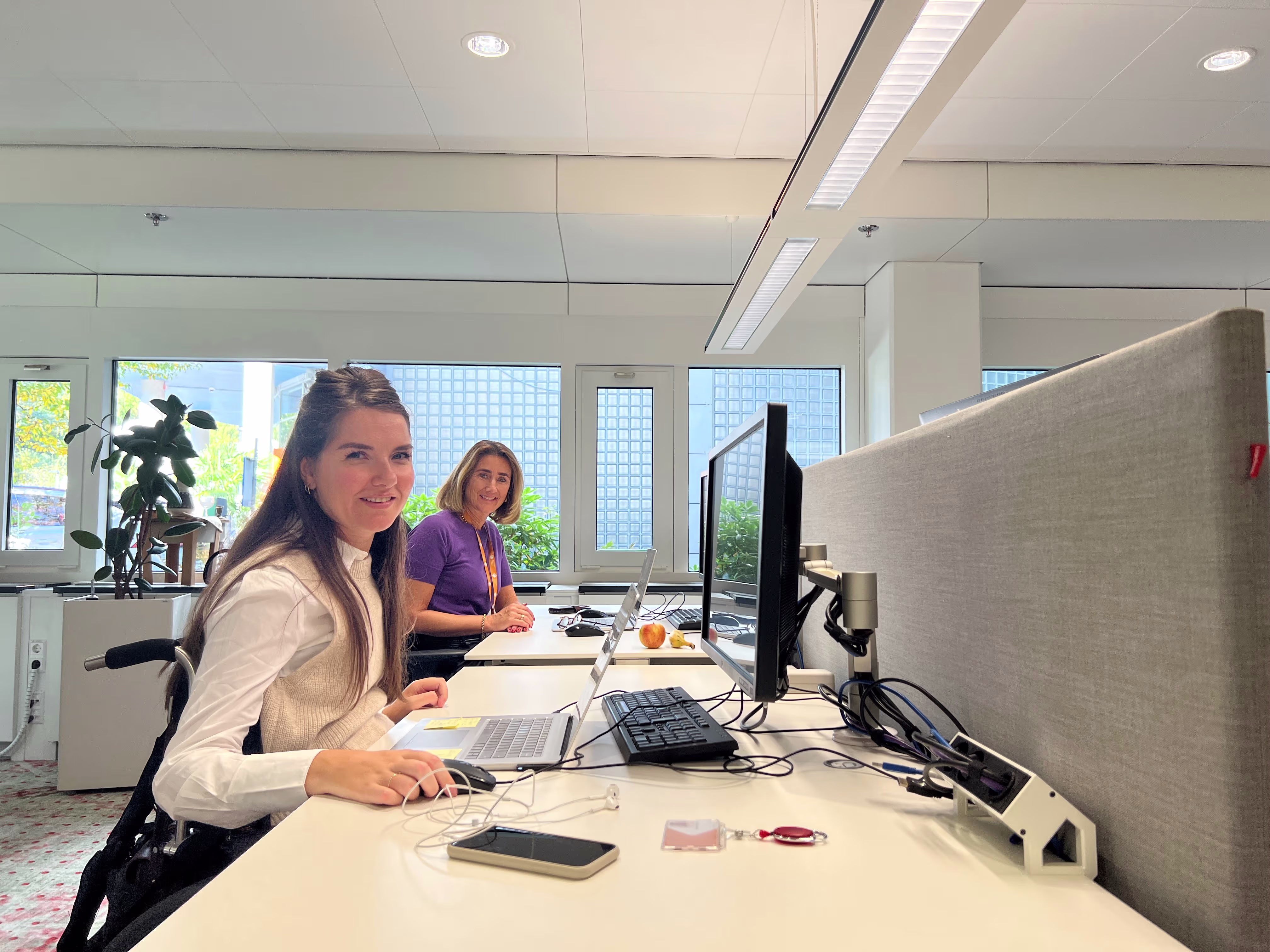 Twee vrouwen werken aan hun bureaus in een moderne kantooromgeving met grote ramen en natuurlijk licht.