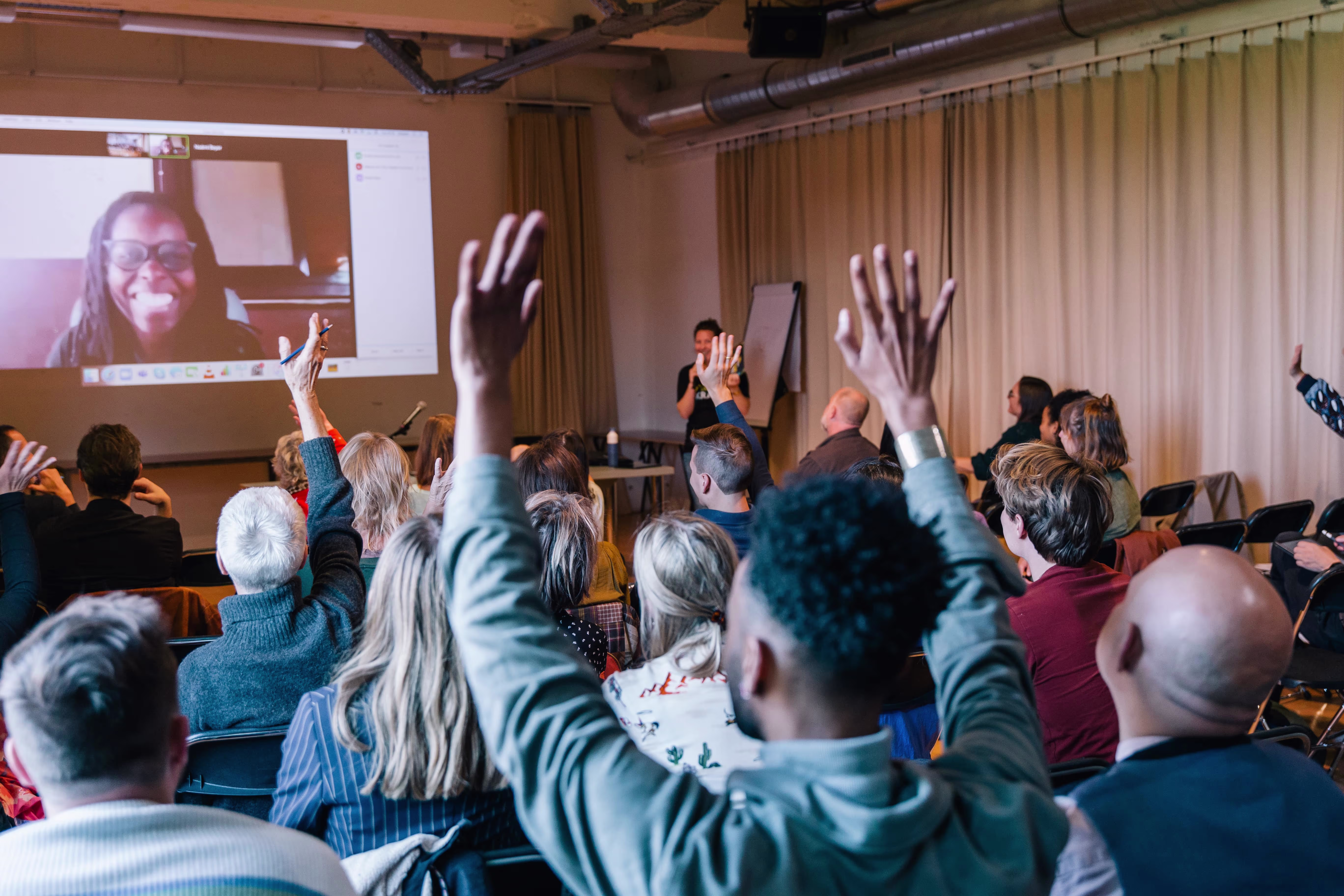 Een groep mensen in een collegezaal steekt hun handen op naar een spreker, terwijl iemand via videoverbinding op het scherm zwaait.
