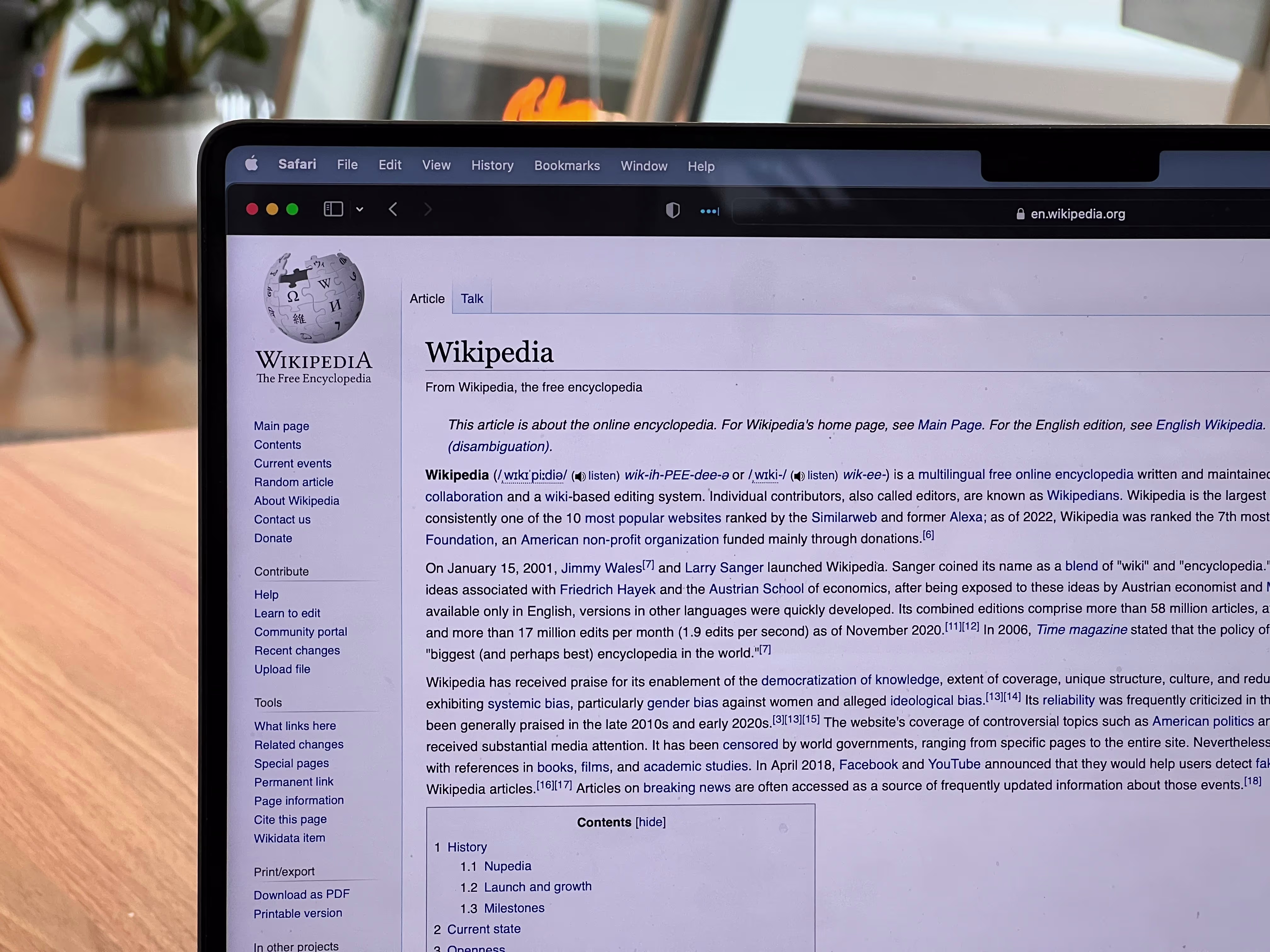 Een laptop toont de Wikipedia-hoofdpagina in Safari browser, op een bureau met huiselijke achtergrond.