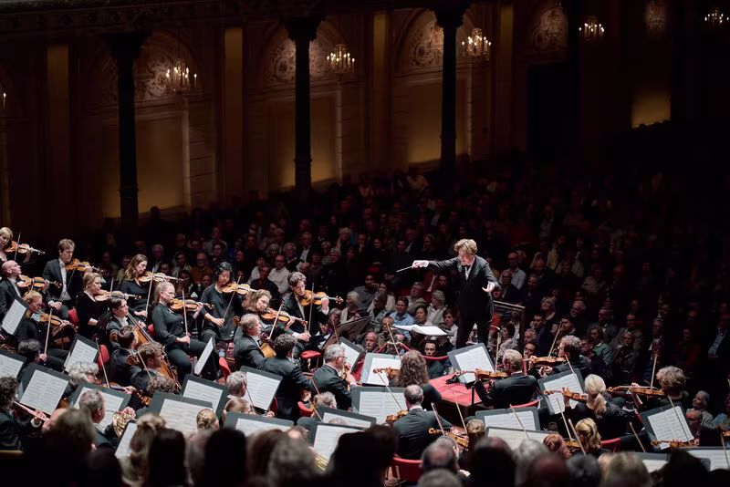Een dirigent leidt een groot orkest tijdens een klassiek concert in een vol concertgebouw met prachtige architectuur.