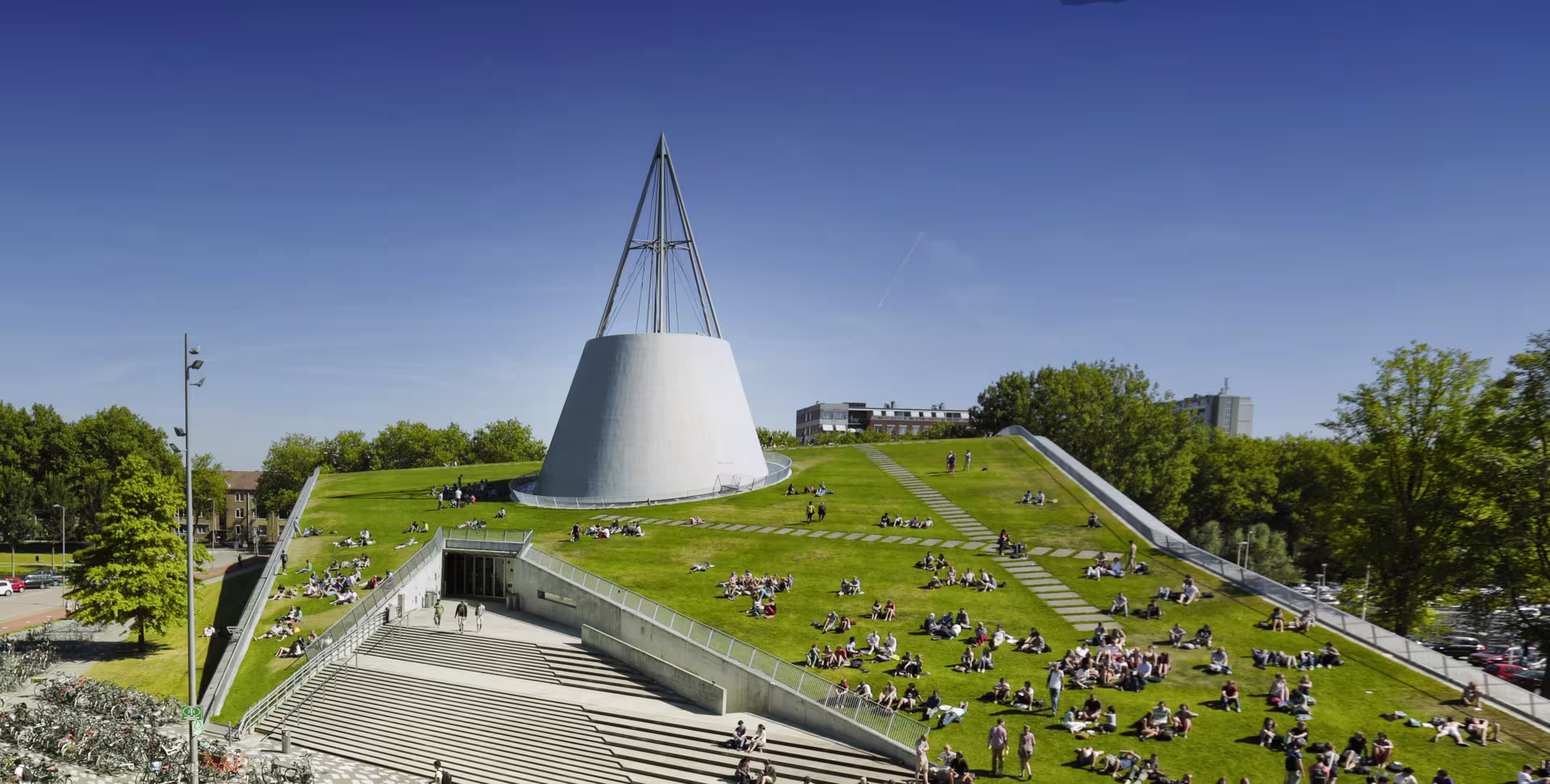 Modern universiteitsgebouw met groen dak waar studenten zitten te studeren onder een blauwe hemel op de TU Delft campus.