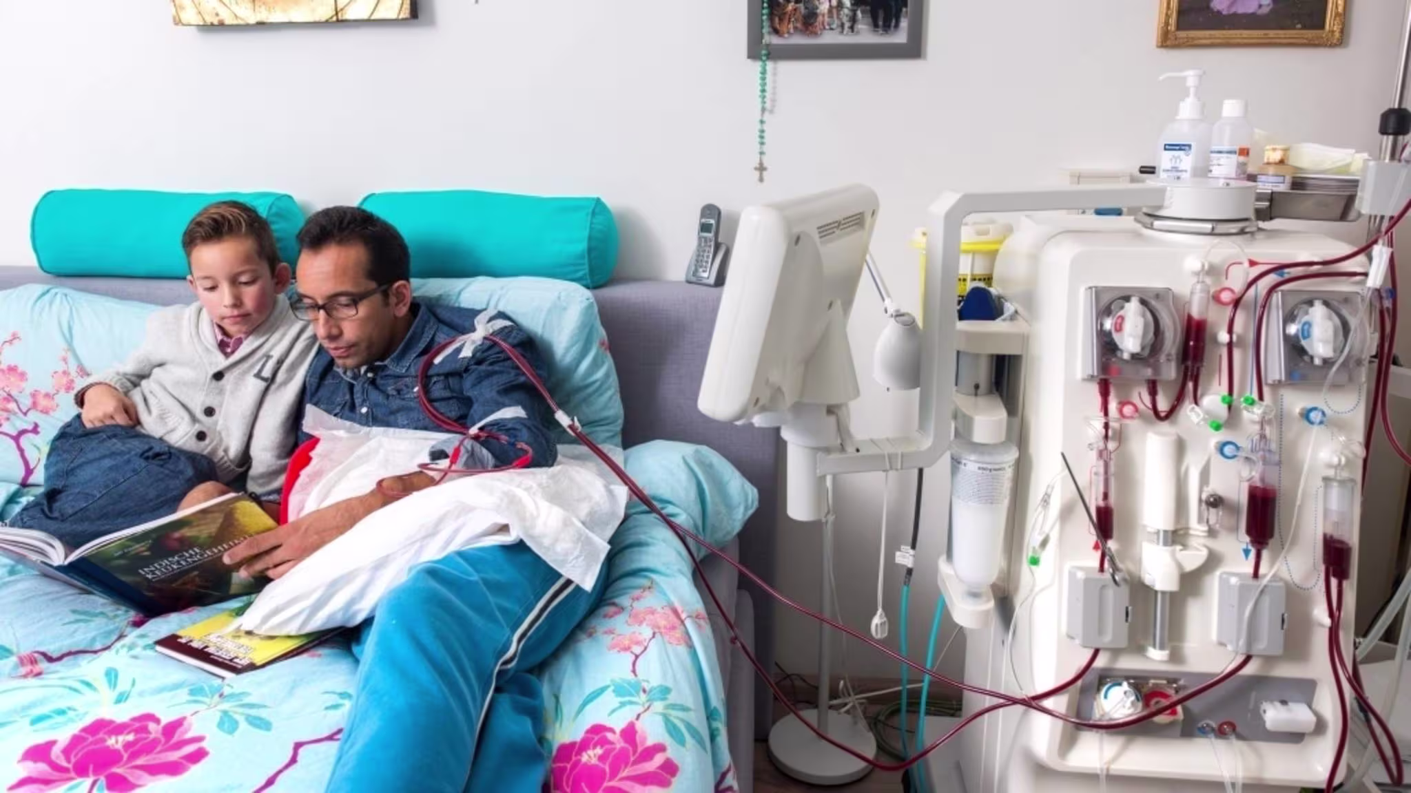 Een vader en zoon lezen samen een boek in een ziekenhuisbed, naast een dialysemachine in een medische kamer.