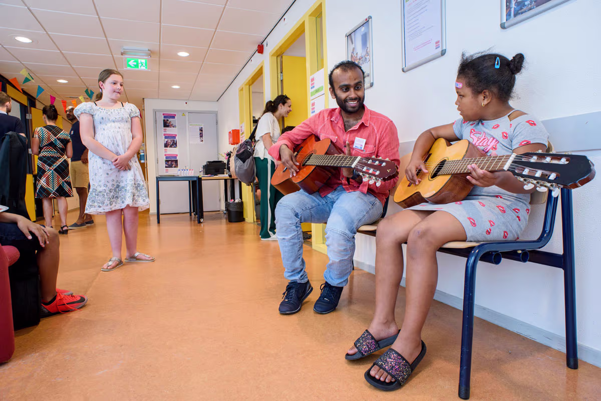 Een muziekdocent speelt gitaar met een leerling in een klaslokaal, terwijl andere kinderen toekijken.