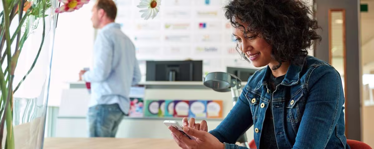 Een vrouw met krullend haar kijkt lachend naar haar telefoon in een modern kantoor. Op de achtergrond werkt een collega.