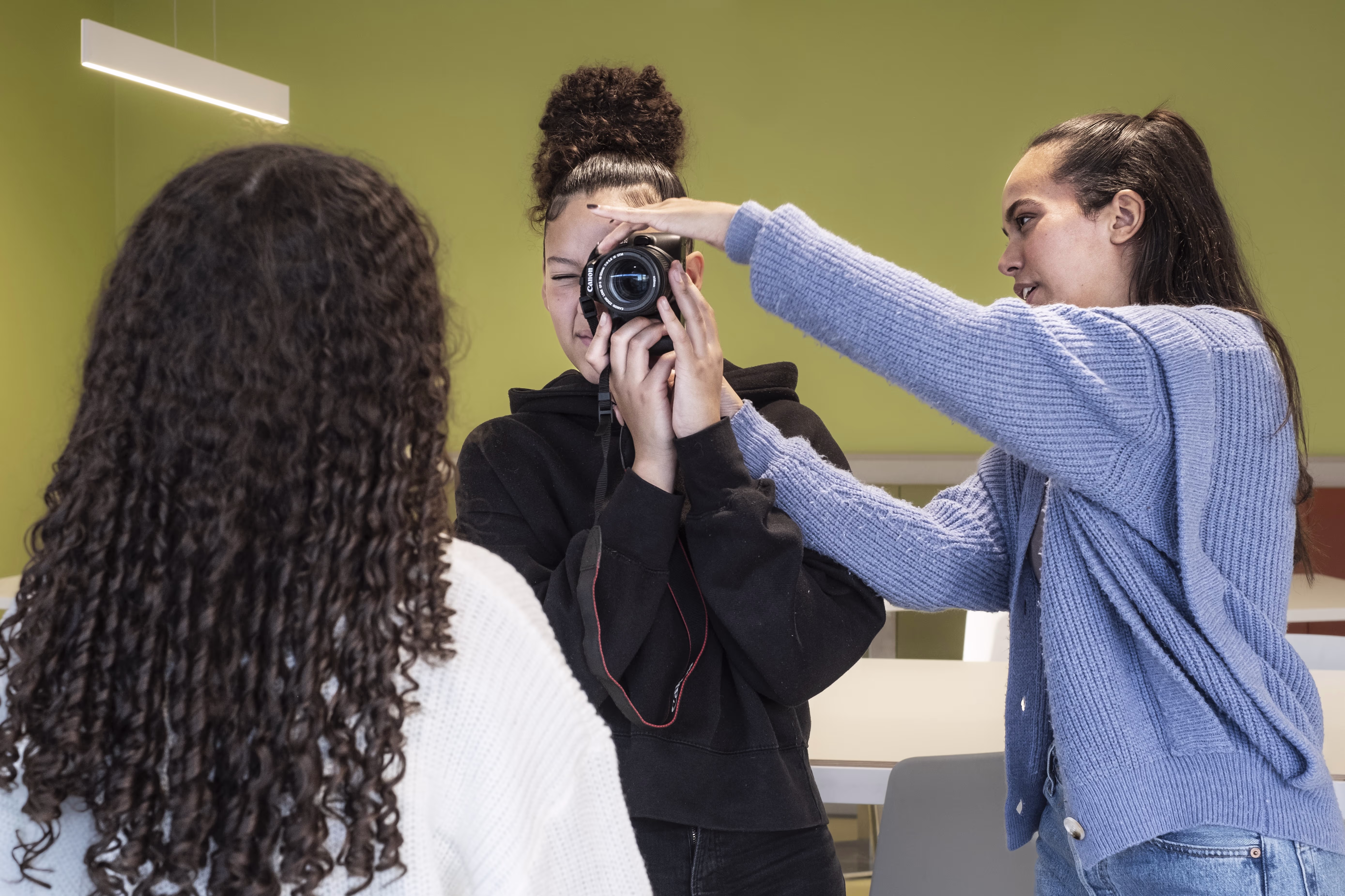 Drie jonge mensen in een kamer, waarvan één een camera vasthoudt terwijl een ander helpt met de belichting of opstelling.