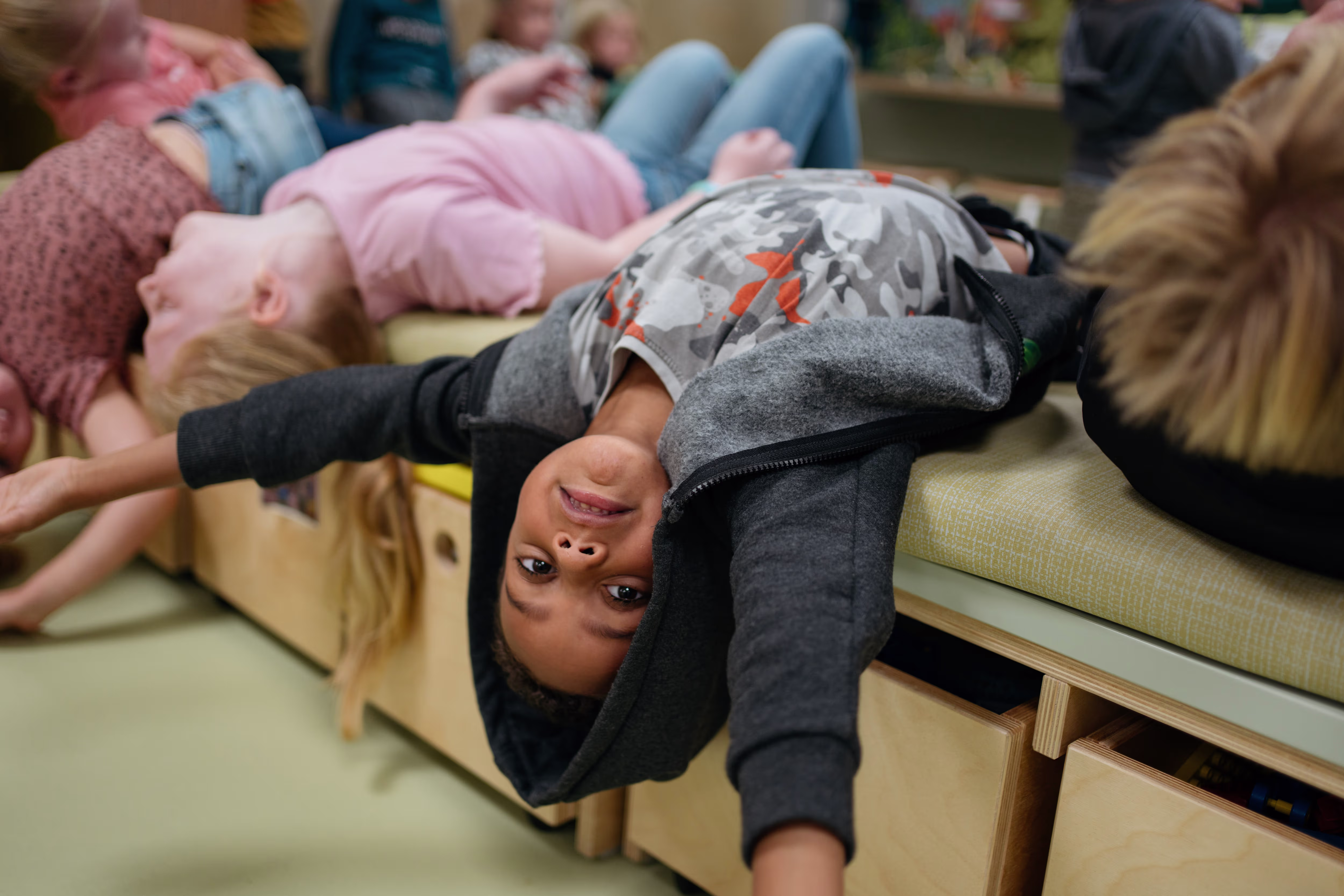 Een kind hangt ondersteboven van een houten bankje en lacht naar de camera, terwijl andere kinderen op de achtergrond spelen.