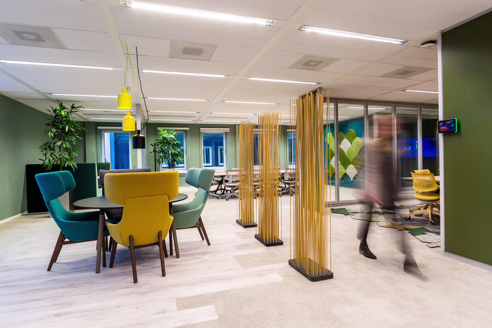 Modern kantoor met gekleurde stoelen rond witte tafels, houten scheidingswanden en glazen vergaderruimtes op de achtergrond.
