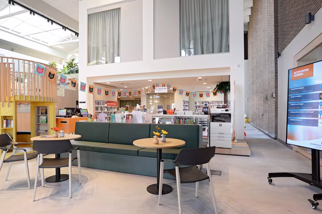 Moderne bibliotheekruimte met groene zitbank, ronde tafels, boeken op de achtergrond en een informatiescherm.