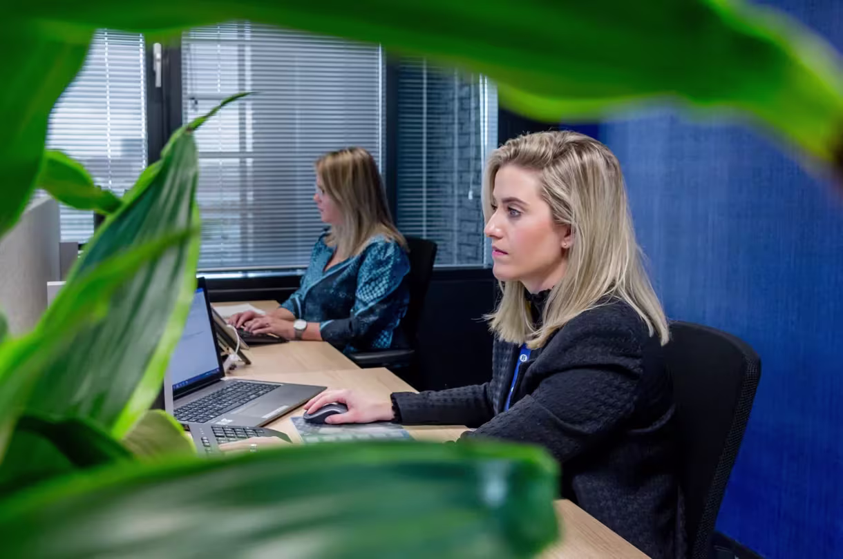 Twee blonde vrouwen werken geconcentreerd op laptops aan een houten bureau in een modern kantoor met groene planten.