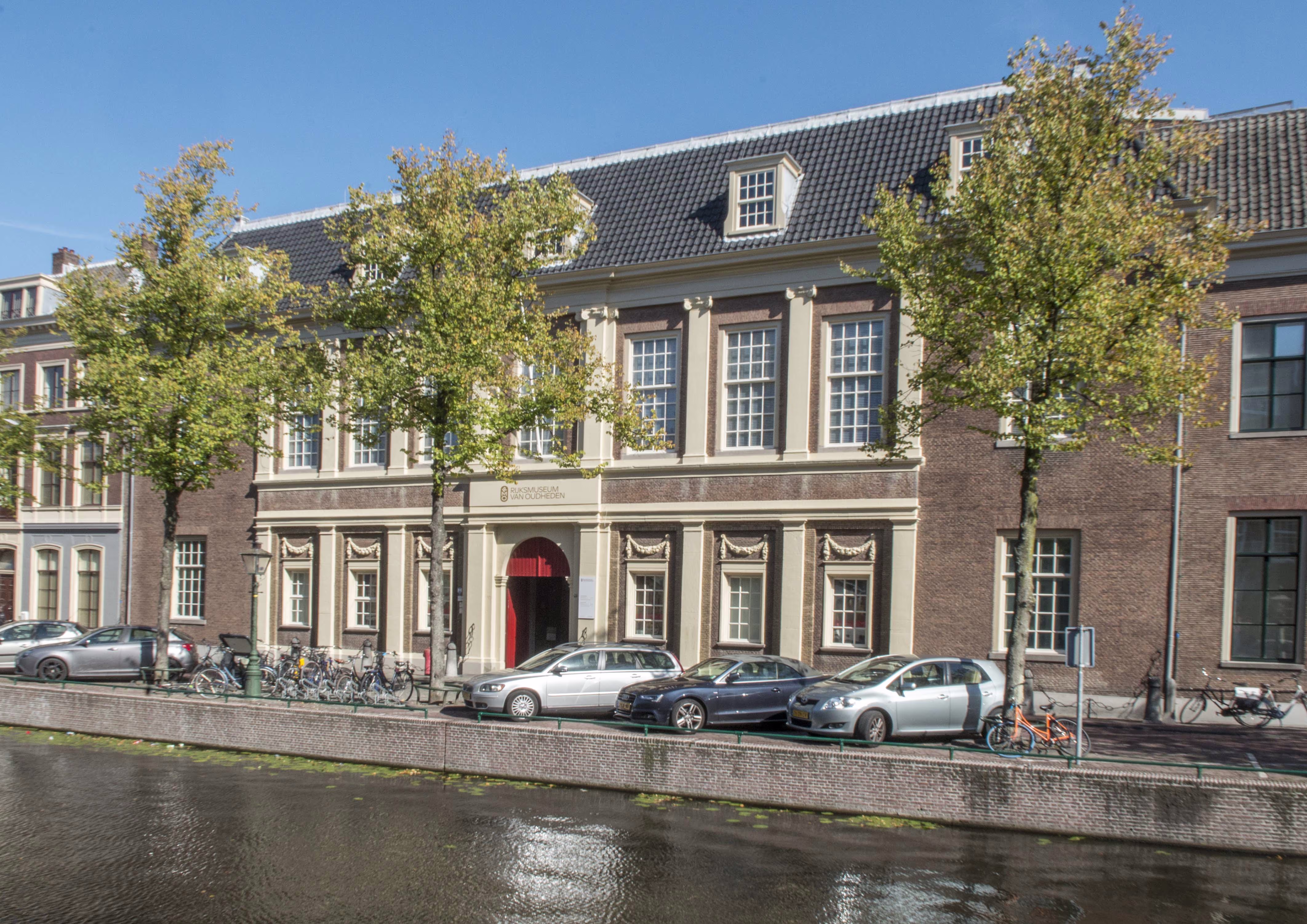 Historisch gebouw van het Rijksmuseum van Oudheden aan een gracht in Leiden met geparkeerde auto's en fietsen ervoor.