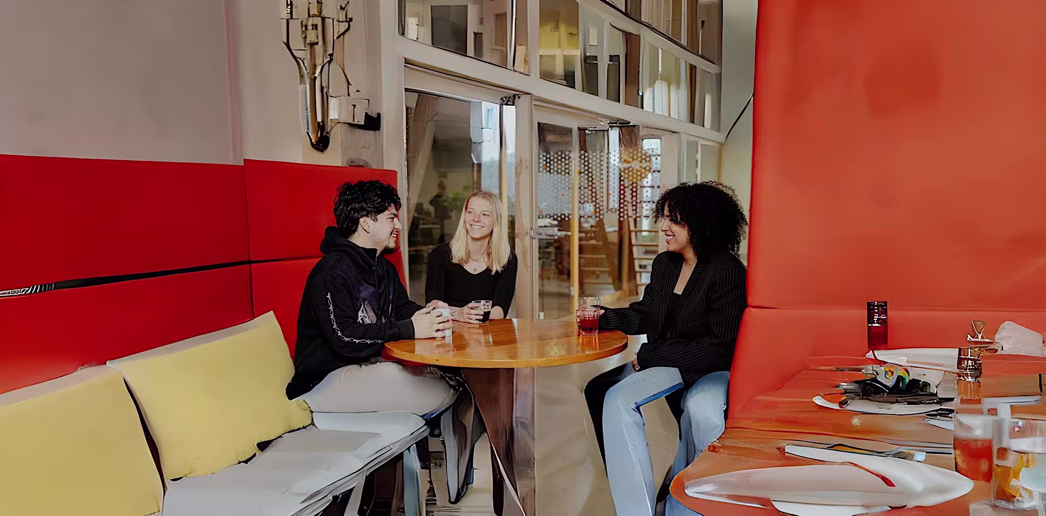 Drie vrouwen zitten aan een ronde tafel in een moderne, oranje-rode kantoorruimte en voeren een gesprek.