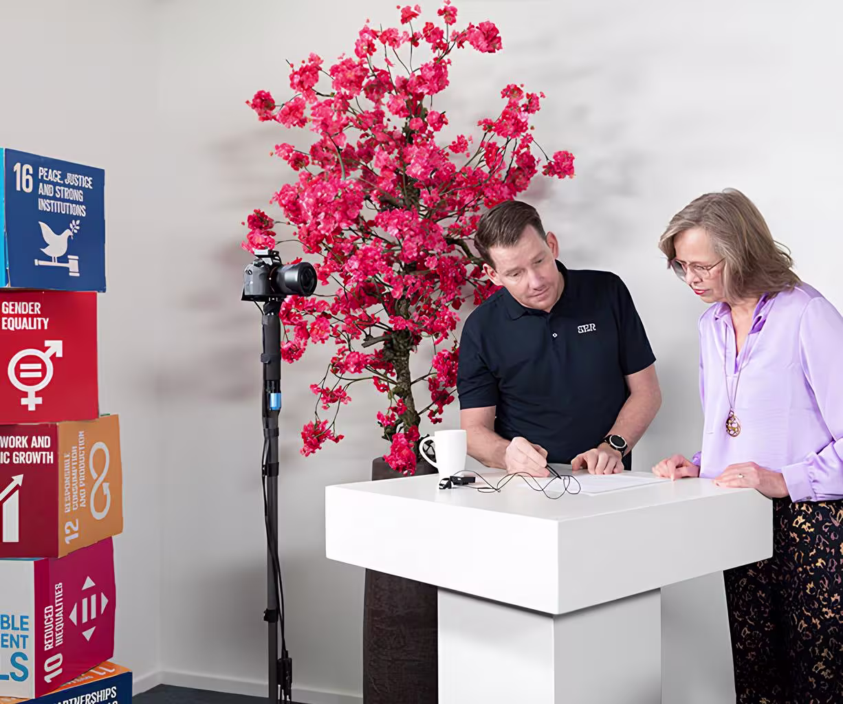 Twee medewerkers bespreken documenten aan een moderne witte tafel, met SDG-poster en roze bloemen op de achtergrond.