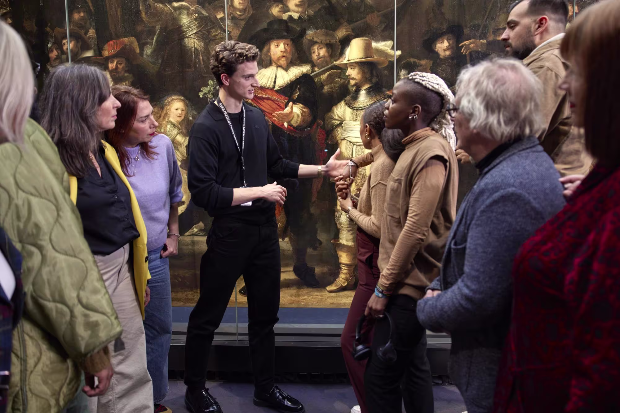 Een museumgids in zwarte kleding legt uit over een klassiek schilderij aan een diverse groep bezoekers in het museum.