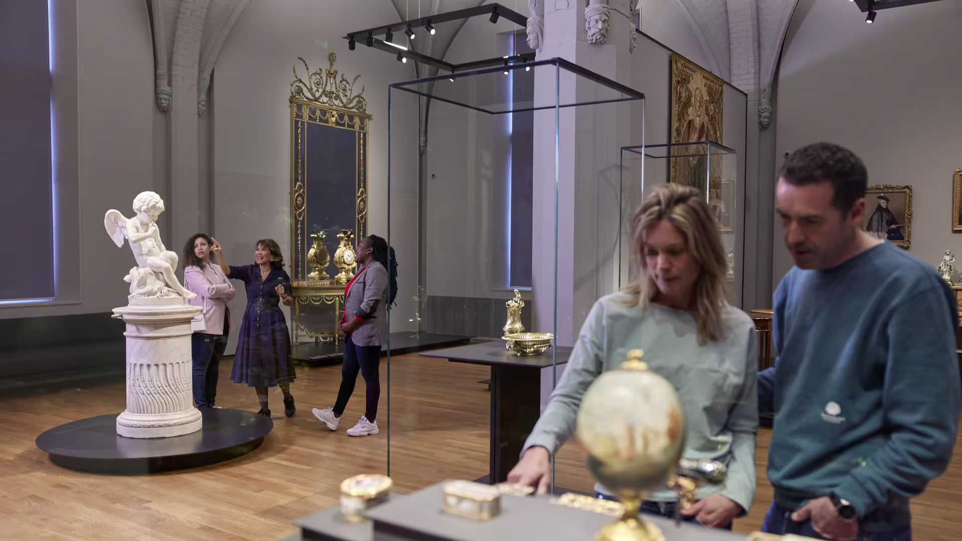 Bezoekers bekijken kunstvoorwerpen in een museumzaal met klassieke sculpturen, schilderijen en een globe in vitrines.