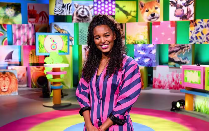 Vrouw in gestreepte blouse staat lachend in kleurrijke tv-studio met wandvullende beeldschermen en diverse afbeeldingen.