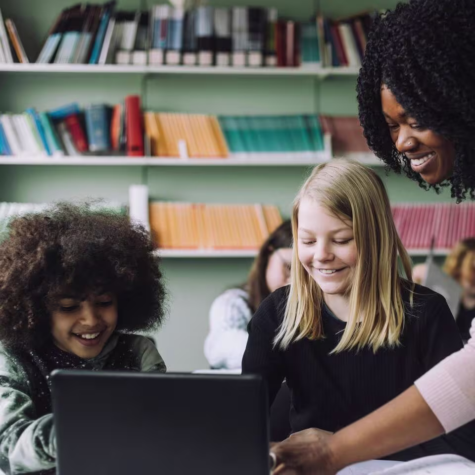 Een vrouw helpt twee kinderen bij een laptop in een bibliotheek met boekenkasten op de achtergrond.