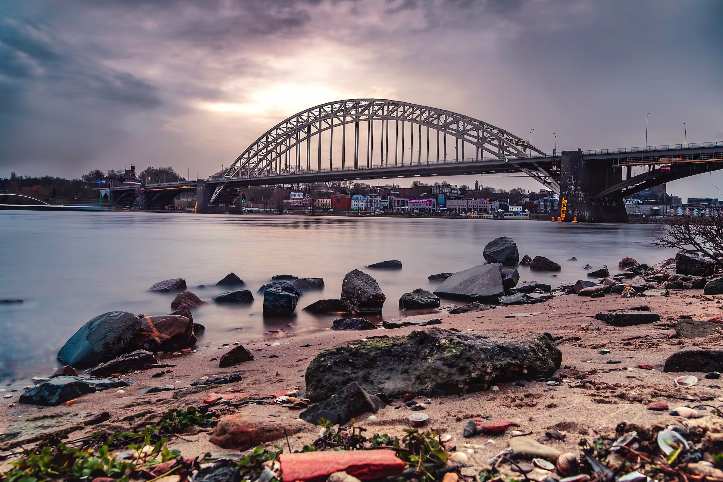Zicht op de Waalbrug in Nijmegen bij zonsondergang, met rotsen langs de waterkant en een dramatische bewolkte lucht.