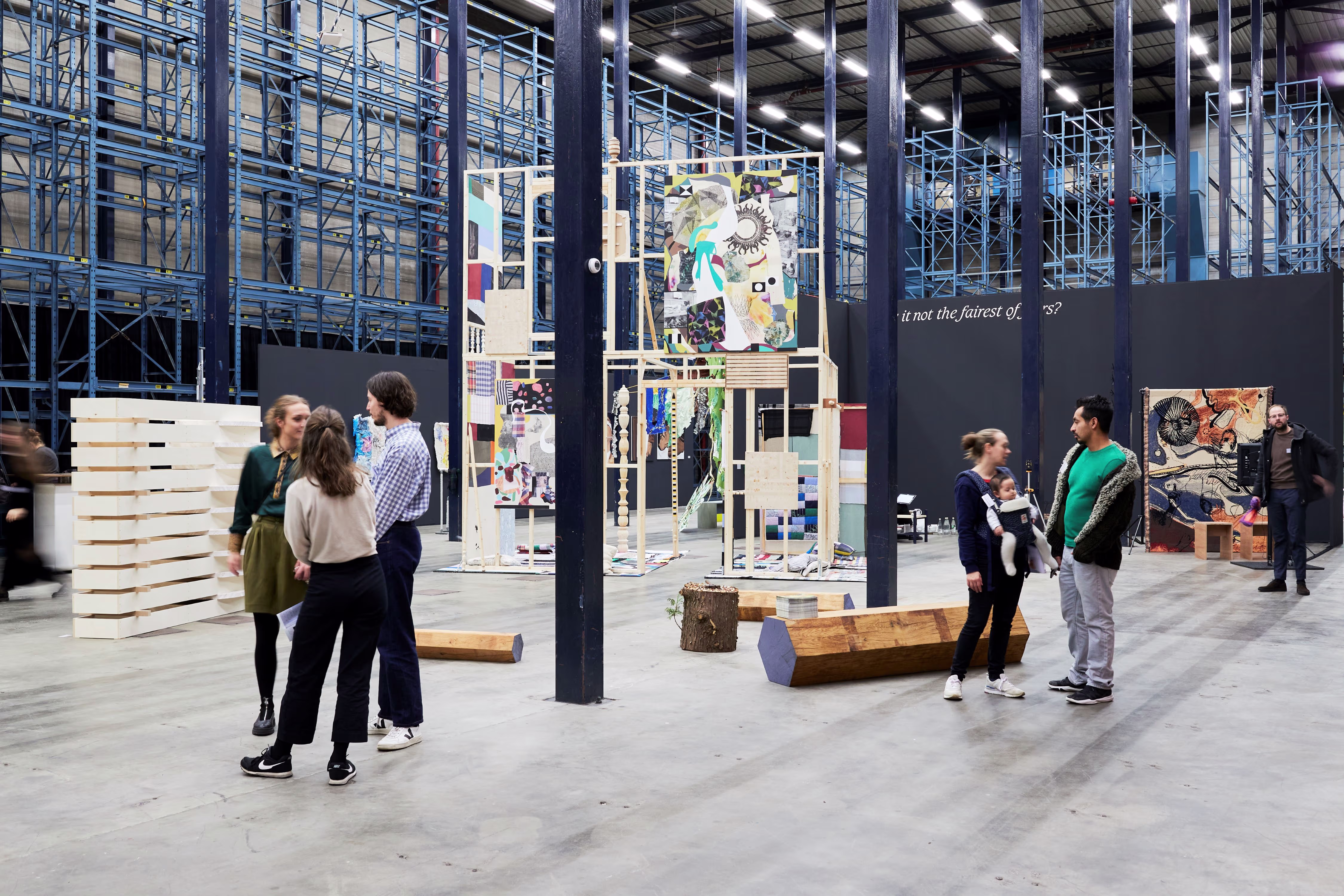Bezoekers bekijken kunstwerken in een grote expositieruimte met industriële uitstraling en blauwe stalen constructies.