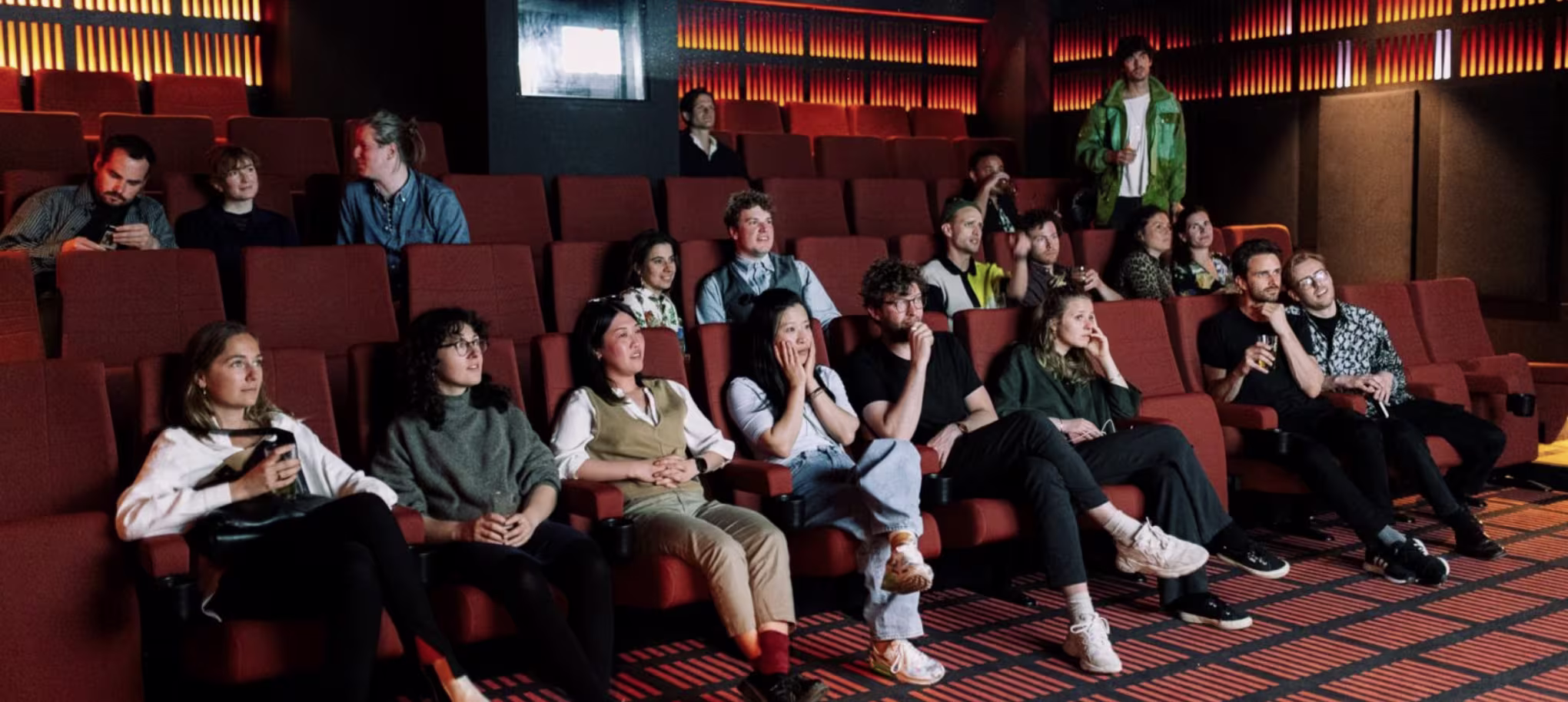 Een groep mensen kijkt naar een film in een bioscoop met rode stoelen en warme sfeerverlichting aan de wanden.