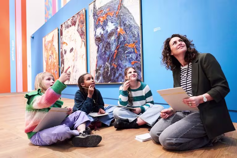 Een museumgids bespreekt schilderijen met drie kinderen die op de grond zitten en naar de kunstwerken kijken.