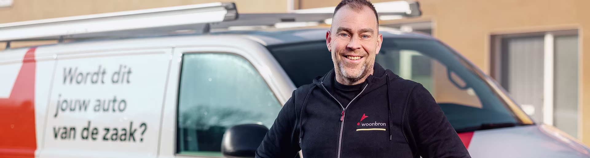 Een lachende man in zwarte werkkleding staat voor een bestelbus met de tekst "Wordt dit jouw auto van de zaak?"