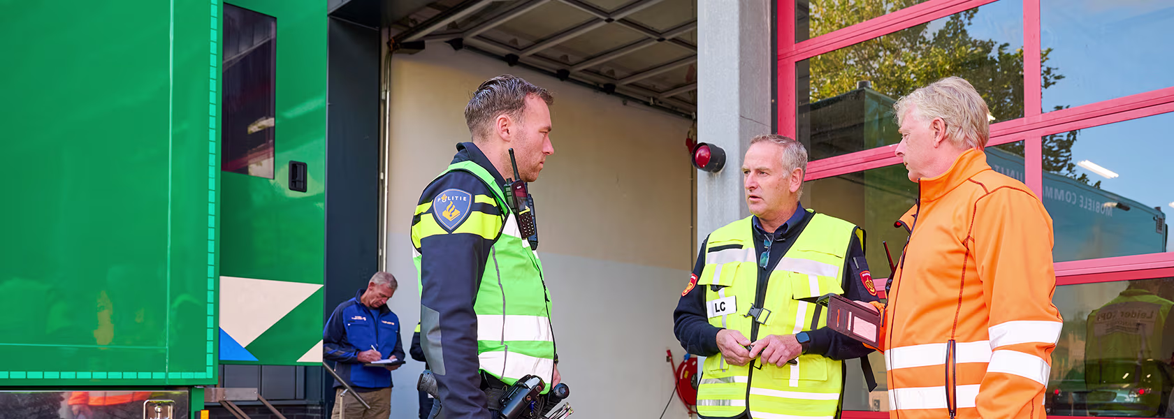 Twee politieagenten in geel-groene hesjes praten met een medewerker in oranje werkkleding bij een brandweerkazerne.