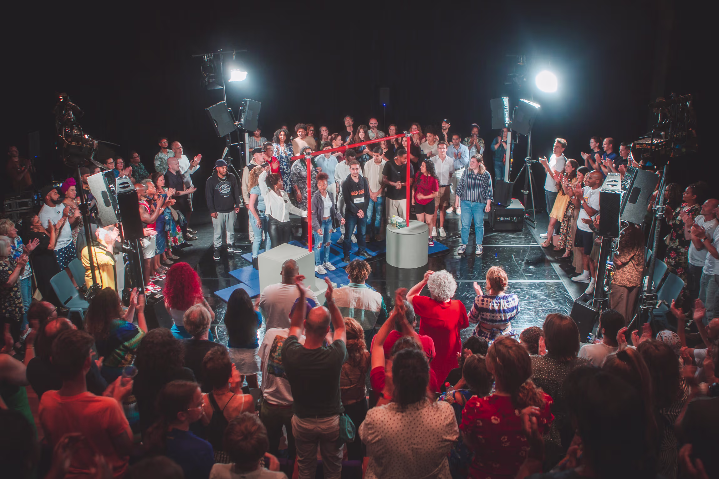 Een grote groep mensen staat in een cirkel rond een podium met professionele filmcamera's en felle studiolampen.