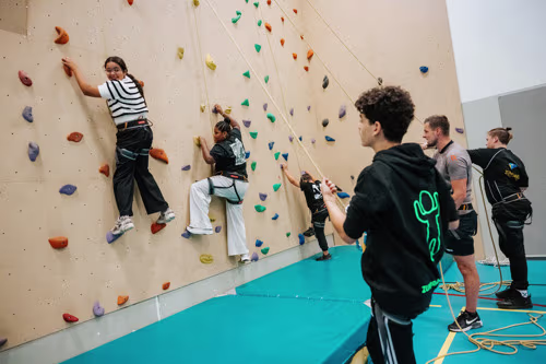 Jongeren klimmen op een indoor klimwand terwijl anderen toekijken in een sporthal met blauwe vloer.
